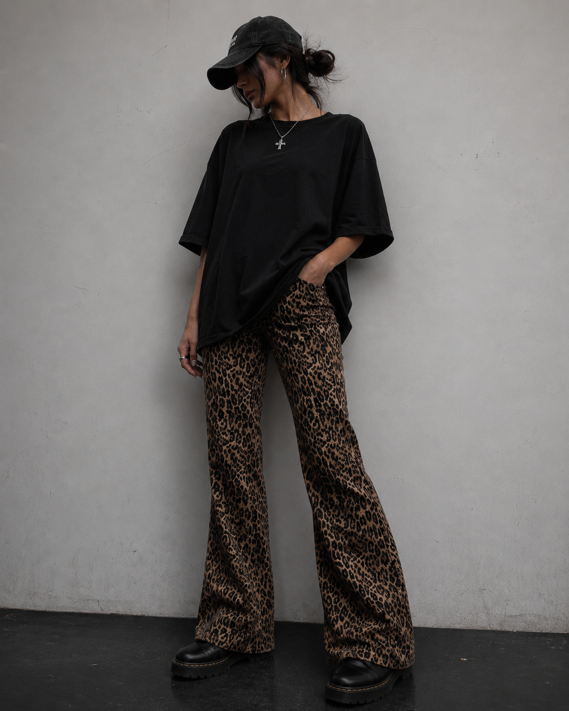 Person wearing a black oversized shirt and leopard print pants against a plain wall.