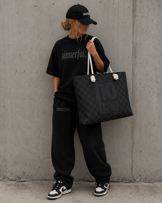 Person wearing black outfit with 'sinnerfold' branding, holding a black tote bag against a concrete wall.