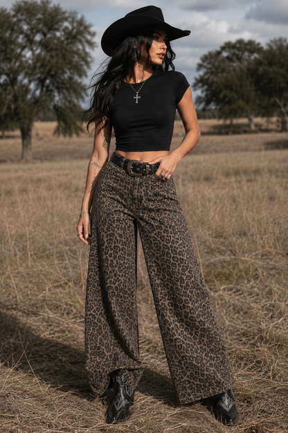 Woman in a black cowboy hat, black top, and leopard print pants standing in a field.